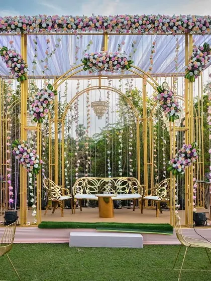 A chic and modern stage setup for a Mehendi or Sangeet. The gold frame structure is decorated with pastel florals and hanging crystals, with a memory wall of photos on the side.