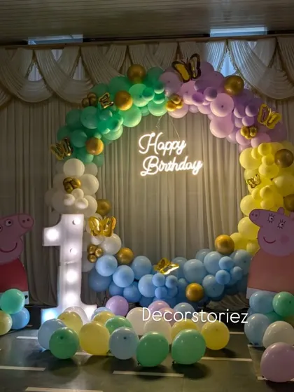 A bright and cheerful Peppa Pig first birthday setup. The colorful balloon ring is accented with golden butterflies and a glowing neon sign for an extra touch of magic.