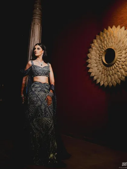 A bride in a dramatic, dark-colored lehenga poses in a dimly lit corridor, her figure illuminated by a single light source.