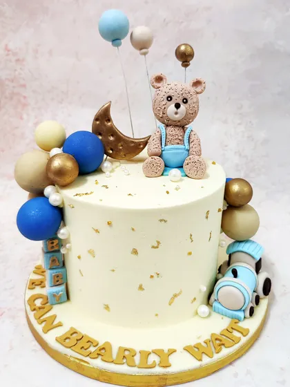 We create a celestial dream with this "We Can Bearly Wait" baby shower cake. A sweet teddy bear sits among stars, a crescent moon, and a toy train on a cream-colored cake accented with blue and gold spheres, perfect for welcoming a new baby boy.
