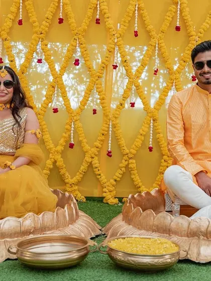The couple seated in large, lotus-shaped brass urlis for their Haldi ceremony, against a backdrop of intricate marigold flower strings.
