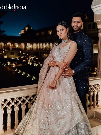 A romantic nighttime portrait of a couple on a balcony at Rambagh Palace, with the illuminated courtyard below.