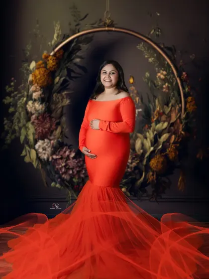 A vibrant and joyful portrait in a stunning orange-red gown. The floral hoop backdrop adds a touch of rustic elegance to this bright and beautiful studio maternity shoot.
