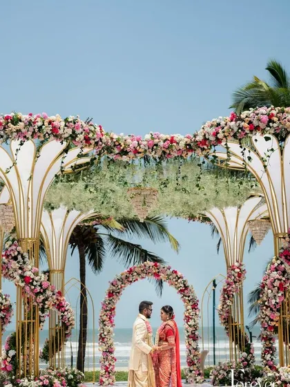 A stunning beachside wedding mandap with intricate white and gold structures, adorned with pink and white flowers.
