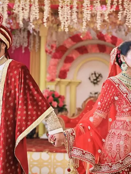 A regal portrait of the couple holding hands, showcasing their coordinated red and white wedding outfits against a decorated backdrop.