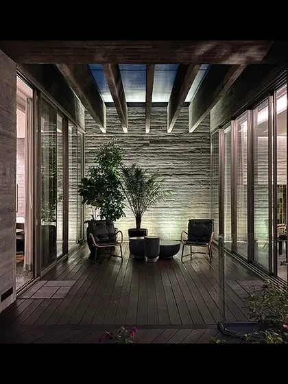 An evening view of a terrace at the Jenga House. The space is framed by exposed concrete walls and wooden ceiling beams, creating an intimate outdoor room that connects directly to the interior.