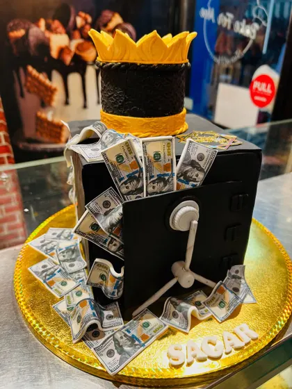 A creative money-themed cake designed as a safe overflowing with edible dollar bills, topped with a golden crown.