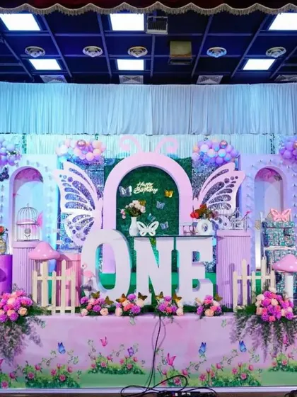 A grand butterfly-themed stage setup. The design features large butterfly wings, flamingo props, and a 'ONE' centerpiece, all in a lovely pink and purple color scheme.