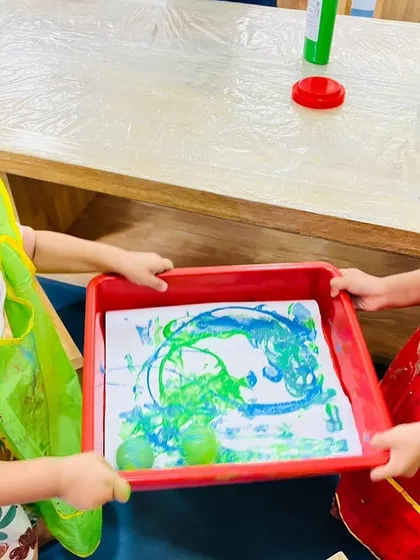 Two children work together, tilting a tray to roll marbles through paint. This activity requires communication and cooperation to guide the marbles and create the art.