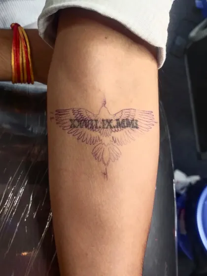Before: A stencil of an eagle is placed over a Roman numeral date tattoo on the forearm, preparing for the cover-up.