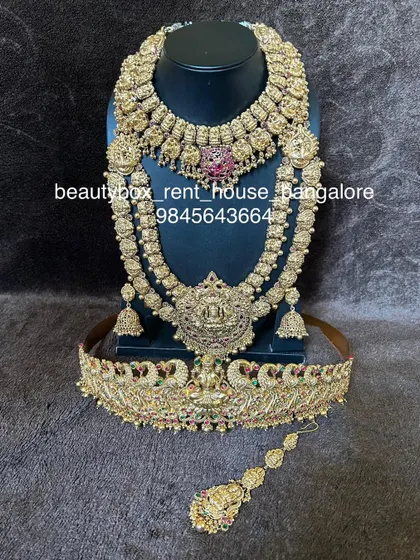A grand bridal set with intricate peacock and Lakshmi motifs, studded with red stones. This rental comes with a choker, long necklace, vaddanam, and maang tikka.