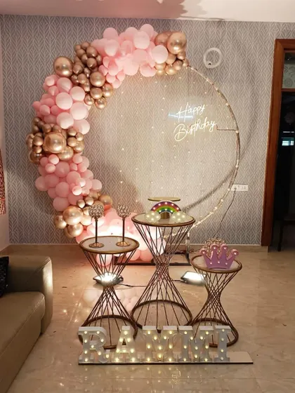 An elegant home decoration featuring a large ring frame with a partial balloon arch in pink and rose gold. The setup is completed with dessert stands and light-up marquee letters.