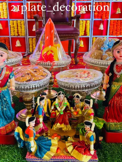 A beautiful and complete baby shower setup with a red and yellow checkered backdrop. The decor includes two large dolls and a central doll scene depicting the Seemantham ceremony.