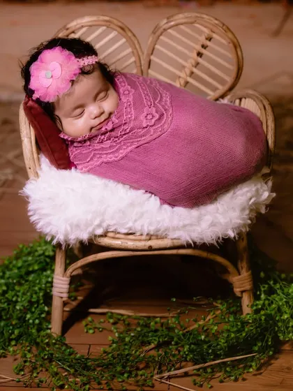 A smiling newborn girl, wrapped snugly in a pink blanket and resting on a miniature cane chair. A truly heartwarming moment.