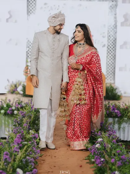 A beautiful couple walking down an aisle lined with purple flowers, against a Sheesh Mahal-inspired backdrop. A picture of timeless elegance.