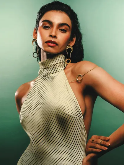 A confident studio portrait for Batoki.art against a rich green backdrop. The model wears a striped halter top and striking gold earrings, showcasing the jewelry in a high-fashion context.