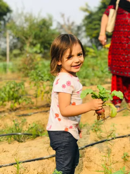 A young girl smiles with delight as she holds up a freshly picked radish. The simple pleasure of harvesting your own food is a core part of the Farmus experience.