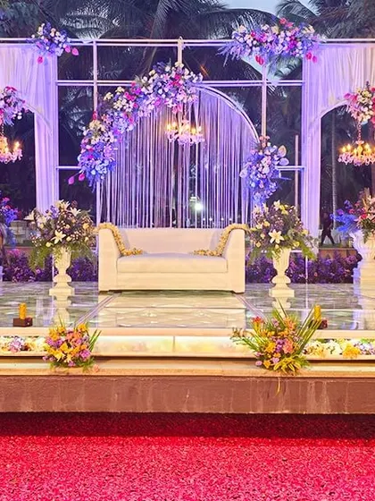 An elegant outdoor reception stage featuring white arched structures, delicate hanging chandeliers, and beautiful floral arrangements. The design is complemented by a floor of scattered red petals, adding a touch of romance.