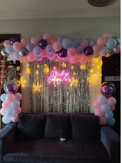 A simple and sweet baby shower decoration designed to fit perfectly in a living room. This setup uses a silver shimmer curtain, a pastel balloon arrangement, and a neon sign placed over the sofa.