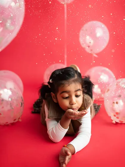 Let's make some magic! A little girl blowing glitter towards the camera against a vibrant red backdrop, surrounded by sparkly balloons. So much fun!