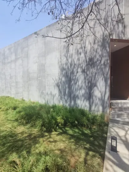 The exterior of a recently completed art gallery and marketing office. The stark concrete wall contrasts with the soft, grassy landscape, while shadows from a nearby tree create dynamic patterns throughout the day.