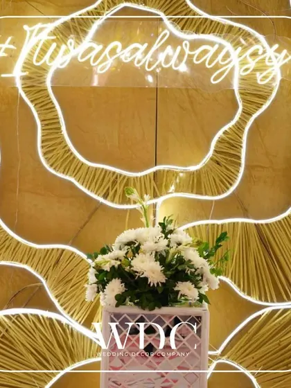 A modern and romantic photo backdrop. A 'It was always you' neon sign is set against a unique, textured golden wall, with a simple floral arrangement in the front.