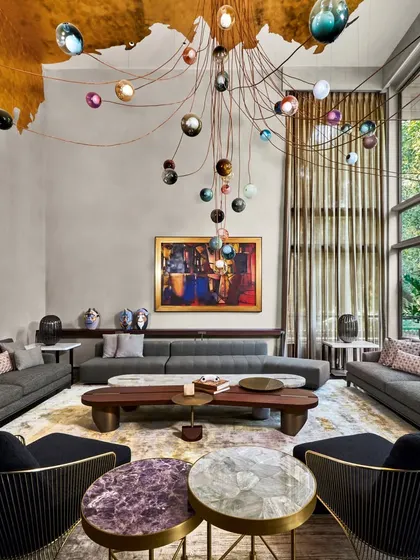 The main living room of the Baid Residence, overlooking lush greens. The space is adorned with modern furnishings, a dazzling Bocci light fixture, a gold leaf ceiling, and captivating artwork by Paresh Maity.