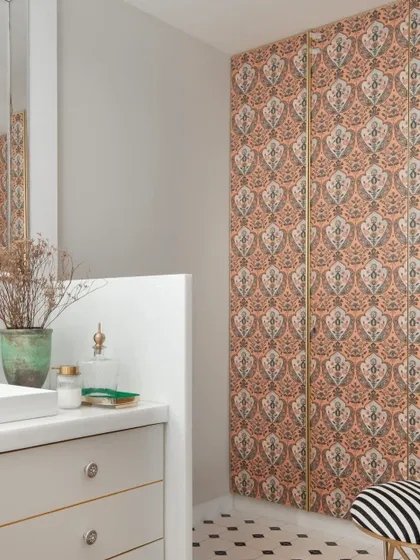 This bathroom features a bold, traditional Indian motif wallpaper in a soft blush tone, creating a statement wall. This is balanced with a simple white vanity and a playful striped stool, mixing patterns with confidence.