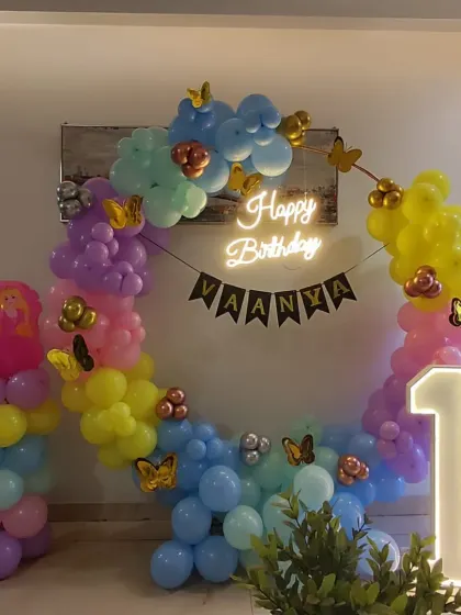 A colorful butterfly-themed first birthday. This decoration uses a multi-color balloon arch, gold butterfly accents, a personalized "VAANYA" banner, a neon sign, and a marquee number '1'.