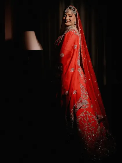 A dramatic bridal portrait using low-key lighting. The single light source illuminates the bride's smiling face and the rich red of her outfit, creating a mysterious and elegant mood.