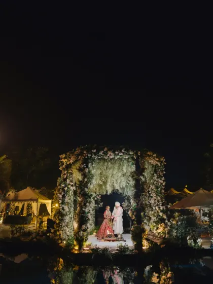 A stunning wide shot of the nighttime ceremony, the floral mandap glowing under the dark sky, its reflection shimmering in the water.