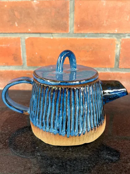 A student's hand-built teapot with a carved vertical line texture and a rich blue glaze. The unglazed base provides a beautiful contrast.