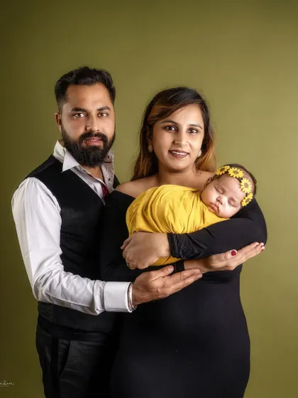 A classic family portrait against a warm, olive green backdrop. The parents hold their newborn daughter together, creating a timeless image of unity and love.