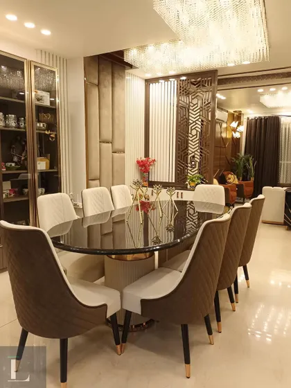A full view of a dining setup in Aradhya Homes, Gurgaon. The oval glass-top table is surrounded by quilted brown and beige chairs, set under a stunning crystal chandelier.