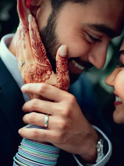 A close-up, intimate moment showing the bride's henna-adorned hand gently touching the groom's face.