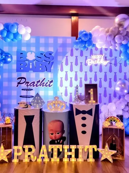 A stylish Boss Baby theme with a checkered blue and white backdrop. The decor includes tie and suit-themed plinths, marquee letters, and a 'Happy Birthday' neon sign.
