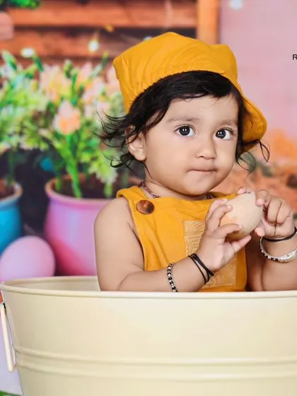 A baby in a yellow hat peeks out of a bucket in this charming bunny-themed garden setting. The curious expression and colorful props make for an adorable photo.
