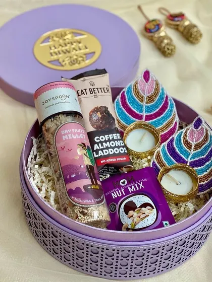 A round rattan box in a lovely lilac color, filled with healthy treats and hand-painted diyas for a unique Diwali gift.