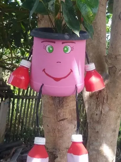This planter hangs from a tree, adding a splash of color and personality to a natural setting. The character is made from a pink container with bottle cap hands and feet.