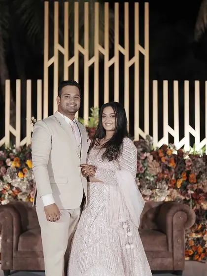A stylish couple poses in front of a contemporary reception backdrop. The geometric white panel and lush floral arrangement create a modern and sophisticated photo opportunity.