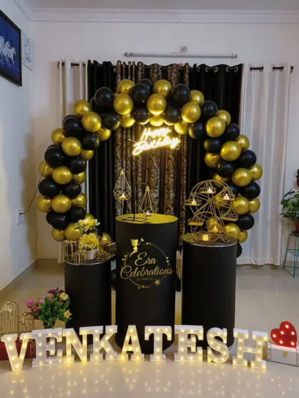 A birthday setup using black plinths and a black and gold balloon arch. The fairy lights and marquee letters create a festive and personalized atmosphere in the home.