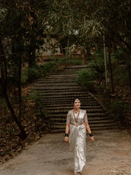 A bride in a traditional white South Indian wedding saree walks down a path in a forested area, creating a beautiful contrast between her attire and the natural surroundings.