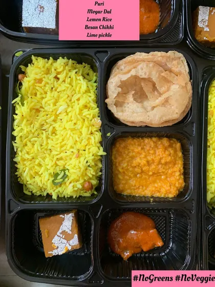 A close-up of a 'No Greens, No Veggies' meal box. This thali includes Puri, Mogar Dal, Lemon Rice, Besan Chikki, and lime pickle, ensuring a complete and tasty meal.
