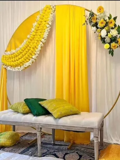 A modern Haldi seating arrangement featuring a circular frame with draped yellow fabric and a crescent of marigold flowers. The elegant bench and cushions complete this stylish look.