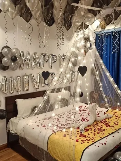 An anniversary room decoration with a fairy light canopy, silver and black balloons, and a swan towel arrangement on the bed.