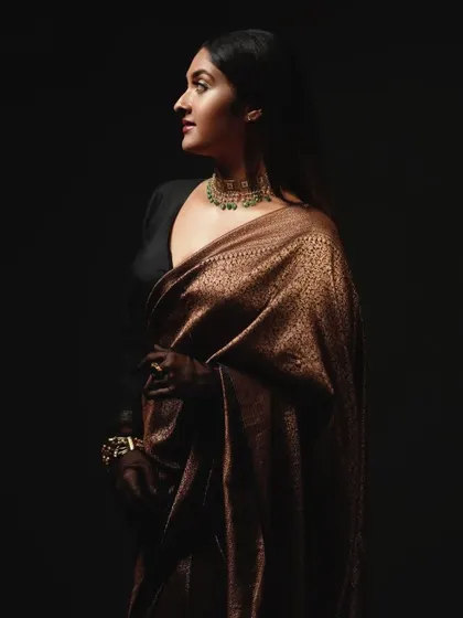 A dramatic, low-key portrait from the 'Raaga' shoot. The lighting highlights the silhouette of the model and the texture of the saree, creating a moody and sophisticated image that focuses on form and shadow.