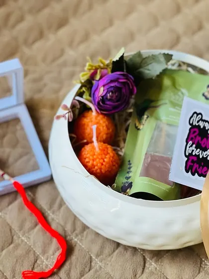 An elegant Rakhi gift set in a white ceramic container with a golden lid, featuring a rakhi and other small, curated items.