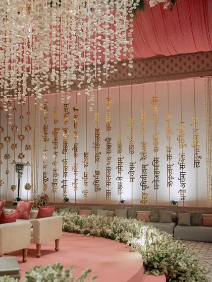 The main ceremony area, featuring a backdrop of hanging golden Sanskrit verses that shimmer in the light. The low seating and surrounding flowers create an intimate and sacred space.