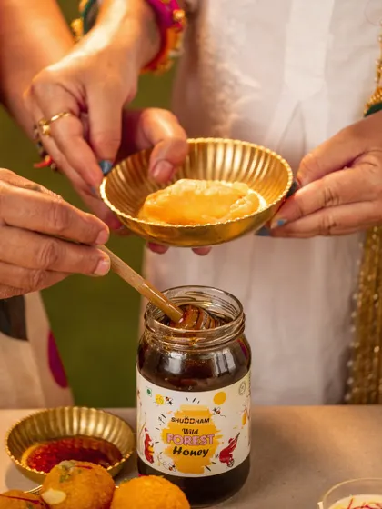 A close-up action shot of hands sharing festive food during a Holi celebration, with a jar of wild forest honey featured prominently.