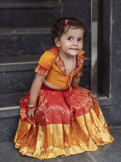 Our cute little client seated on the stairs, looking adorable in her custom orange and red brocade Langa Dhawani.